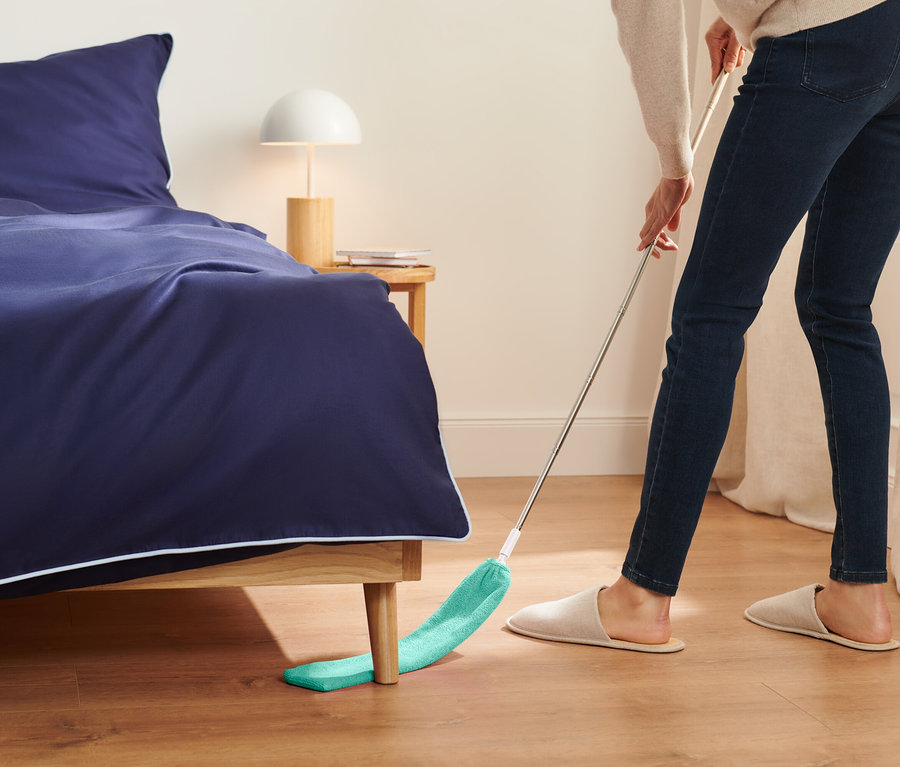 Kobieta zamiata podłogę pod łóżkiem zielonym mopem.