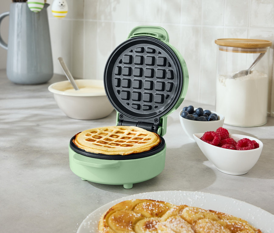 Otwarta, okrągła, miętowo-zielona gofrownica z gofrem w środku stoi na blacie kuchennym. Obok znajdują się dwa spiralne wypieki na talerzu, miska z jagodami i miska z malinami.