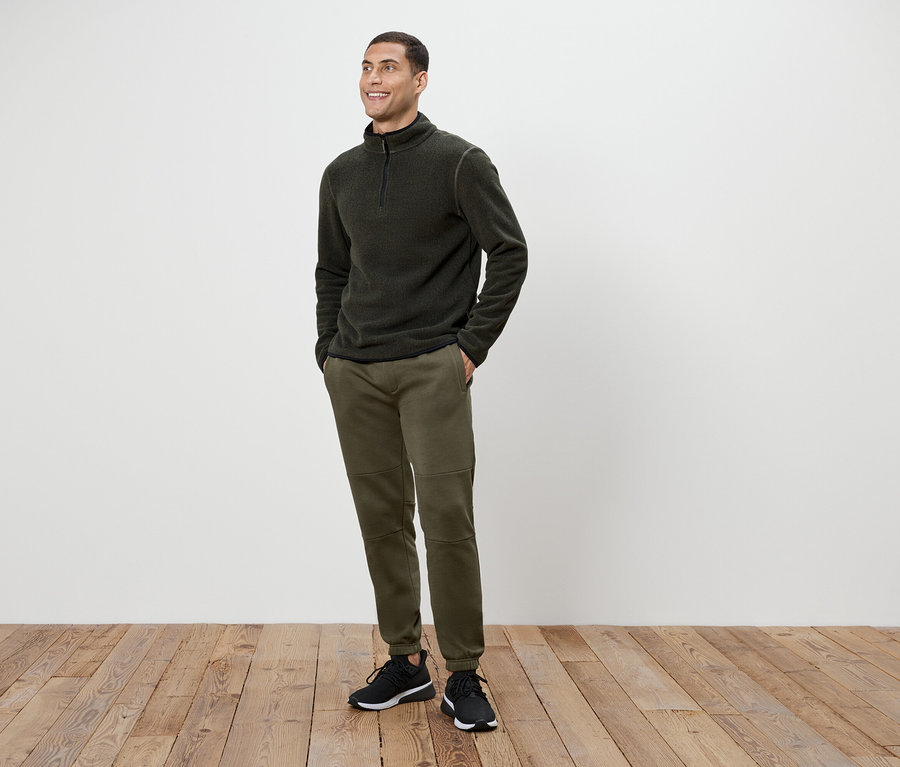 Mężczyzna w oliwkowych spodniach i ciemnoszarej bluzie polarowej na zamek stoi na drewnianej podłodze.