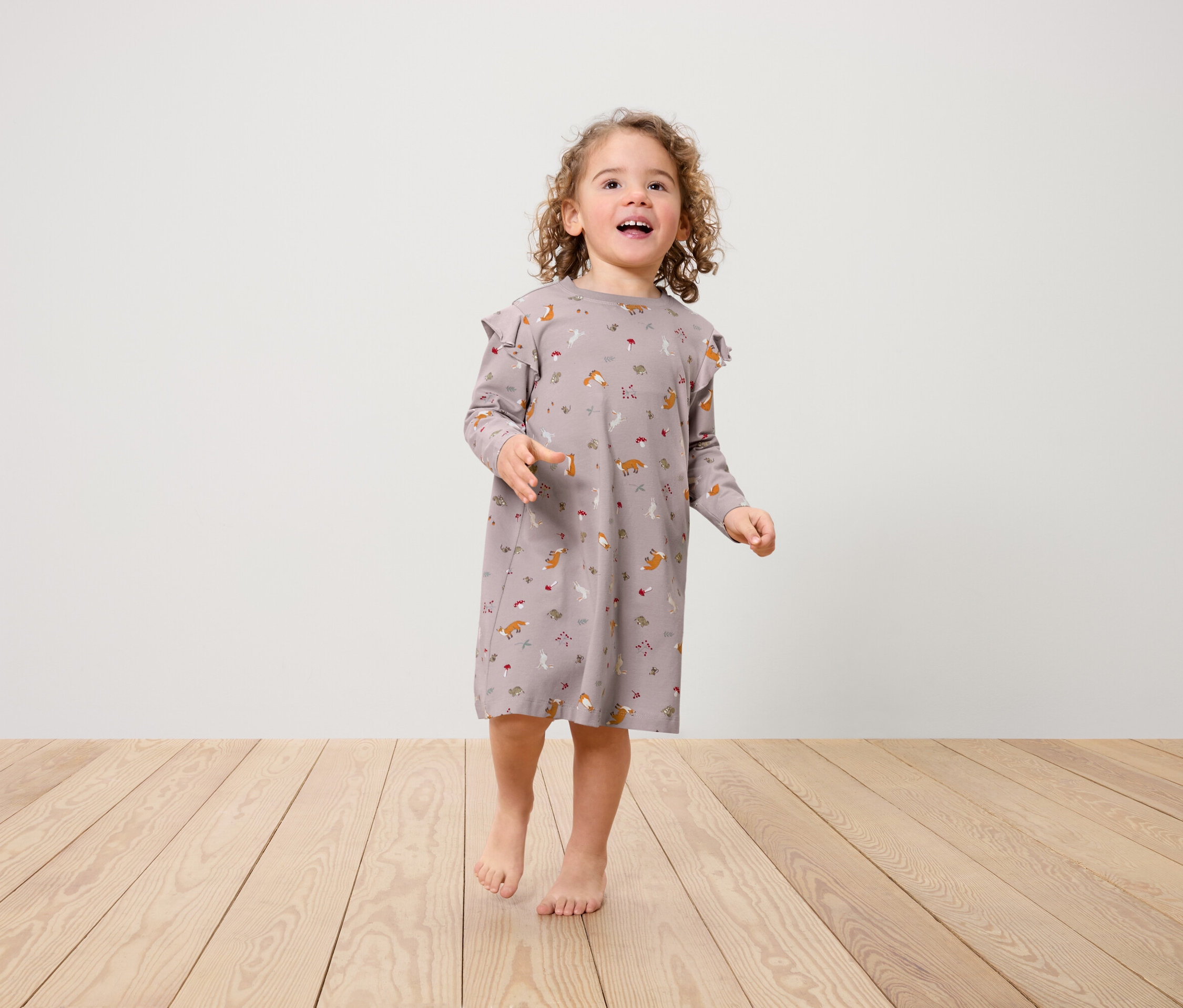 Dziewczynka w sukience w lisy z falbankami na ramionach stoi na drewnianej podłodze.