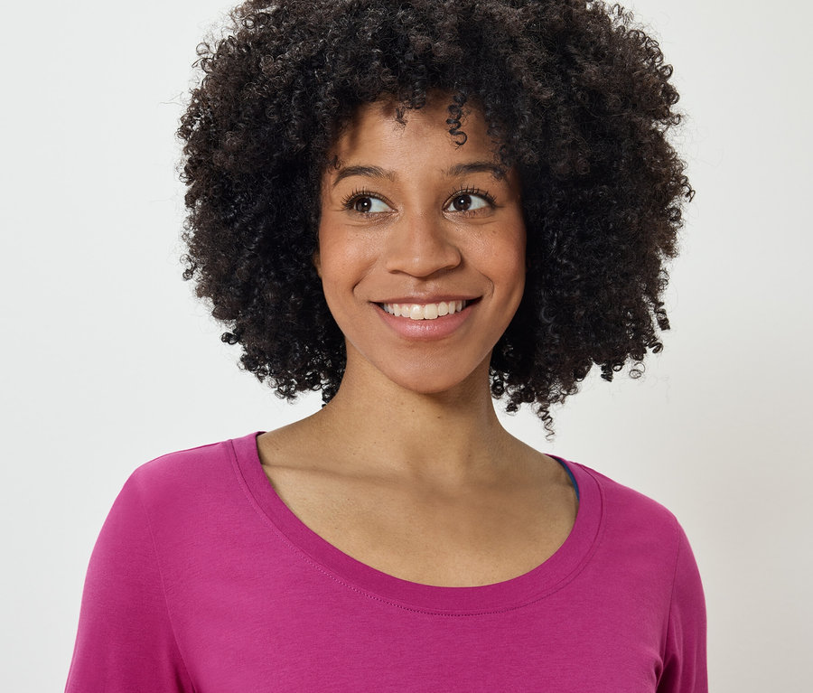 Portret uśmiechniętej kobiety z fryzurą afro w różowej koszulce, patrzącej w lewo.