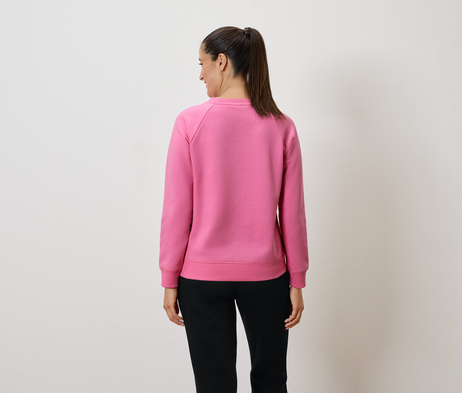 Kobieta w różowej bluzie i czarnych spodniach stoi tyłem do aparatu.