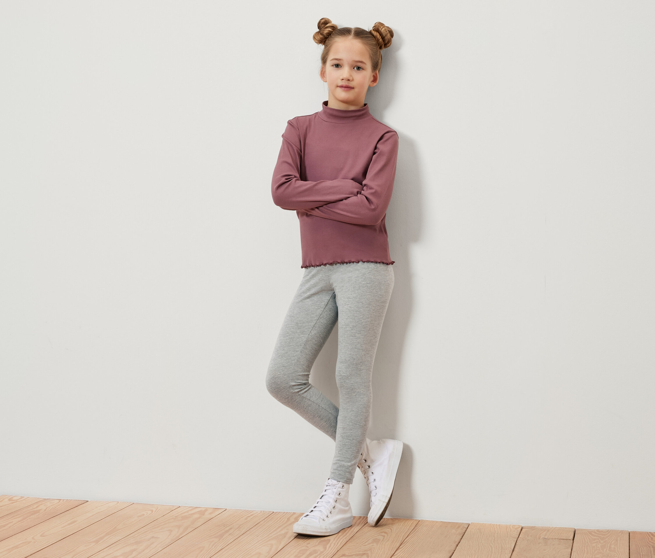 Dziewczynka z brązowymi włosami spiętymi w koczki stoi ze skrzyżowanymi ramionami. Ma na sobie bordową bluzkę z długim rękawem, szare legginsy i białe tenisówki.