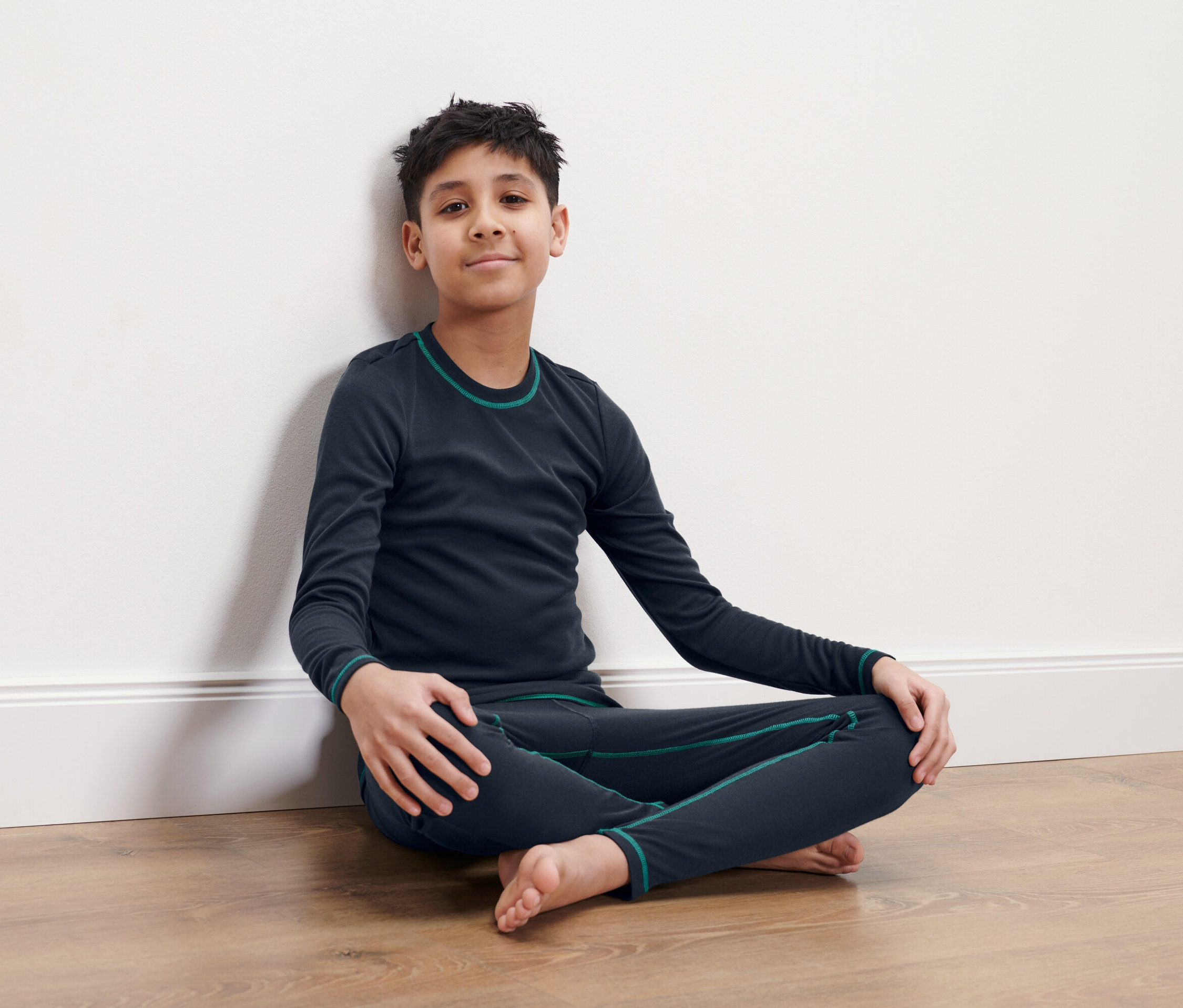 Chłopiec siedzi ze skrzyżowanymi nogami na podłodze, opierając się o białą ścianę. Ma na sobie granatową bieliznę termiczną.