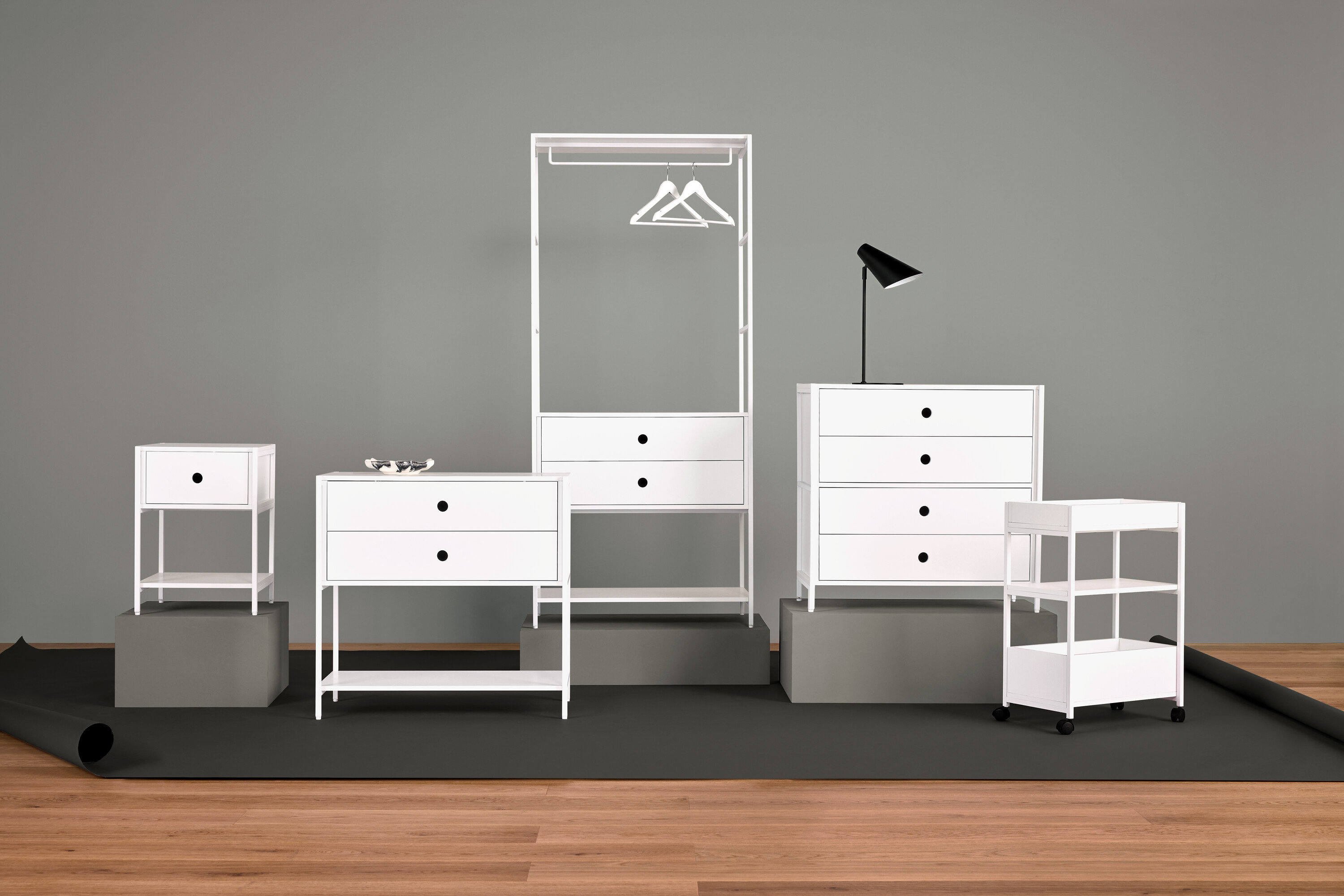 Aranżacja białych mebli z szafką nocną, komodą z szufladami, wieszakiem na ubrania, lampą i półkami.