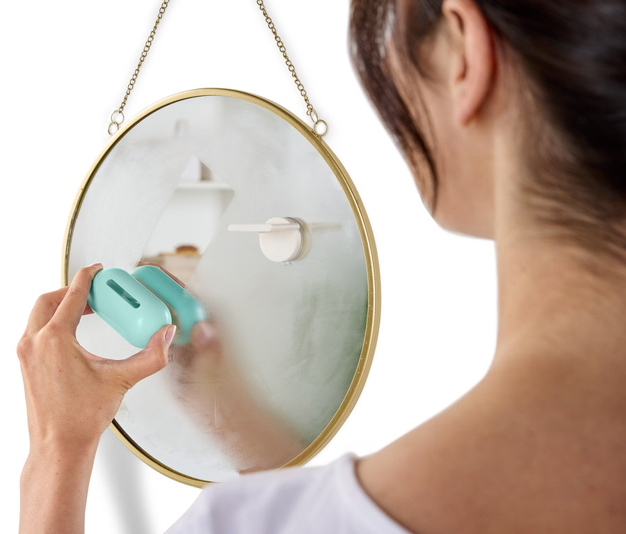 Kobieta czyści okrągłe lustro wiszące na ścianie za pomocą niebieskiego środka czyszczącego.