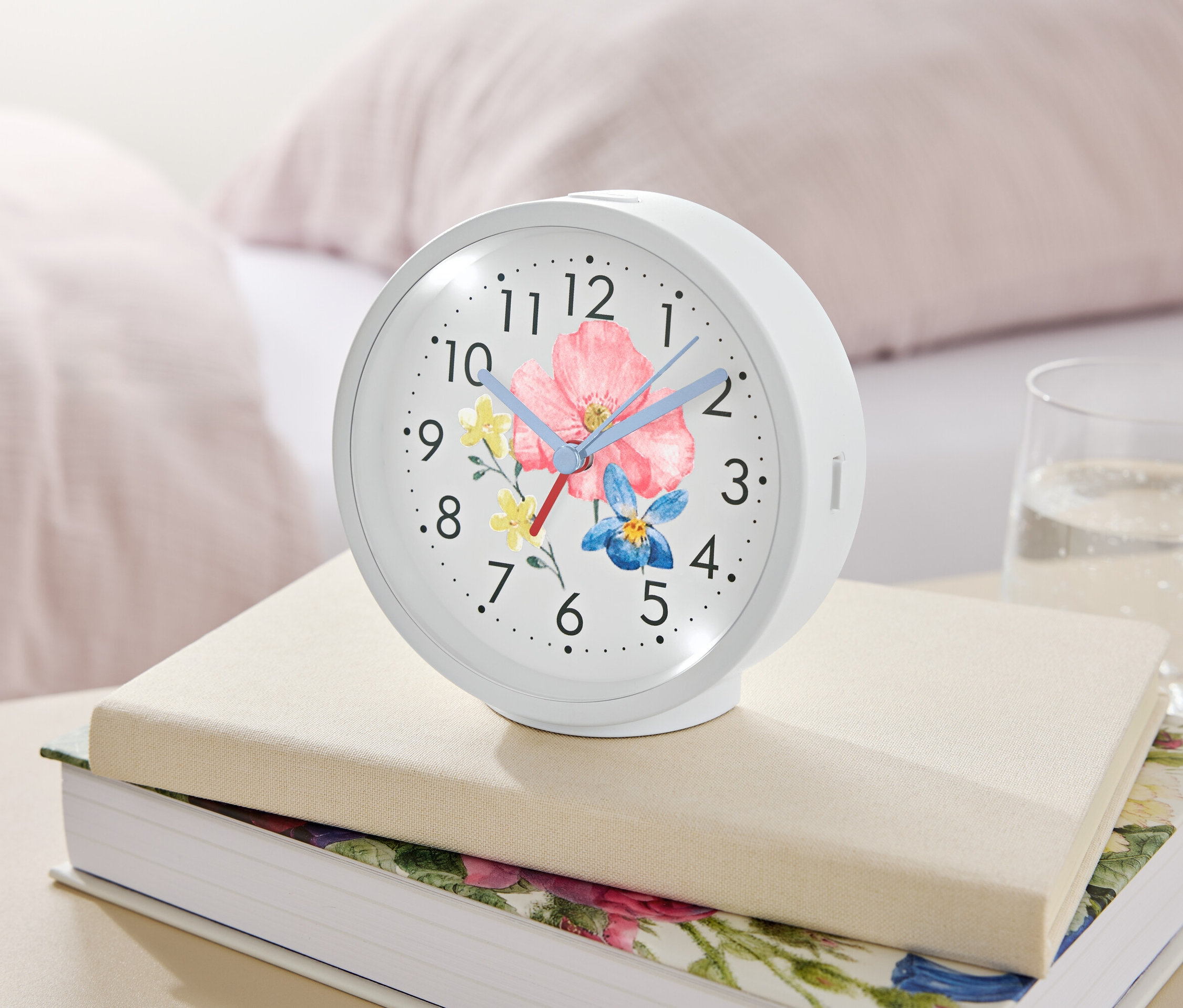 Biały budzik z kwiatowym motywem leży na książkach obok szklanki wody.