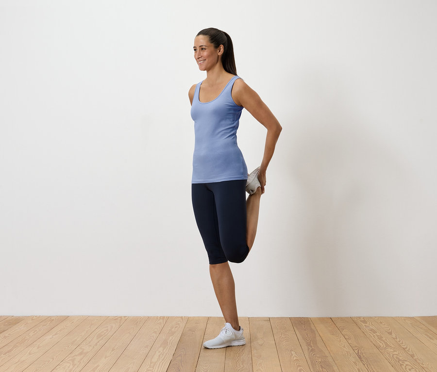 Kobieta w niebieskim podkoszulku i ciemnych legginsach rozciąga mięsień czworogłowy, trzymając stopę za sobą.