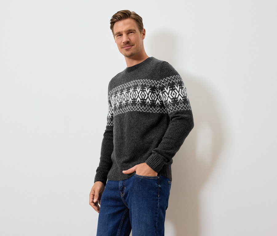 Mężczyzna pozuje w szarym swetrze we wzory i niebieskich dżinsach.