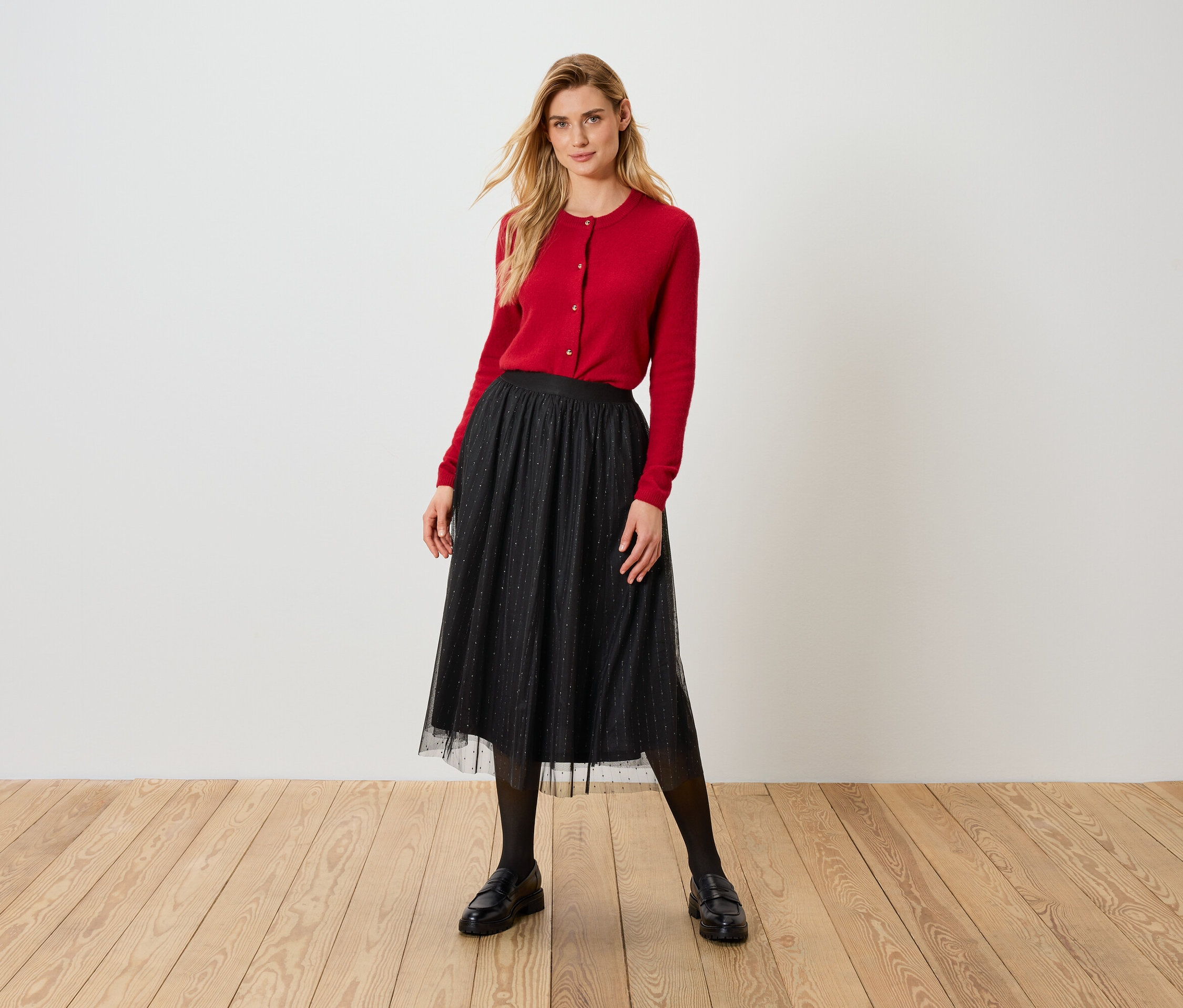 Blondynka w czerwonym kardiganie i czarnej tiulowej spódnicy stoi na drewnianej podłodze.