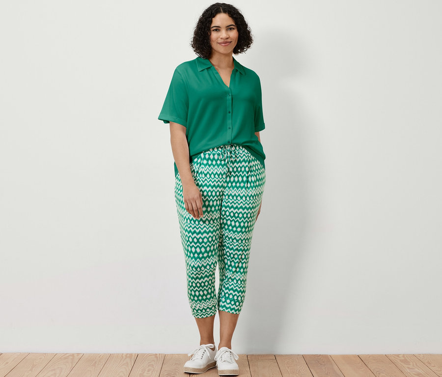 Kobieta stoi ubrana w szmaragdową bluzkę zapinaną na guziki i wzorzyste spodnie dżersejowe o długości 3/4.