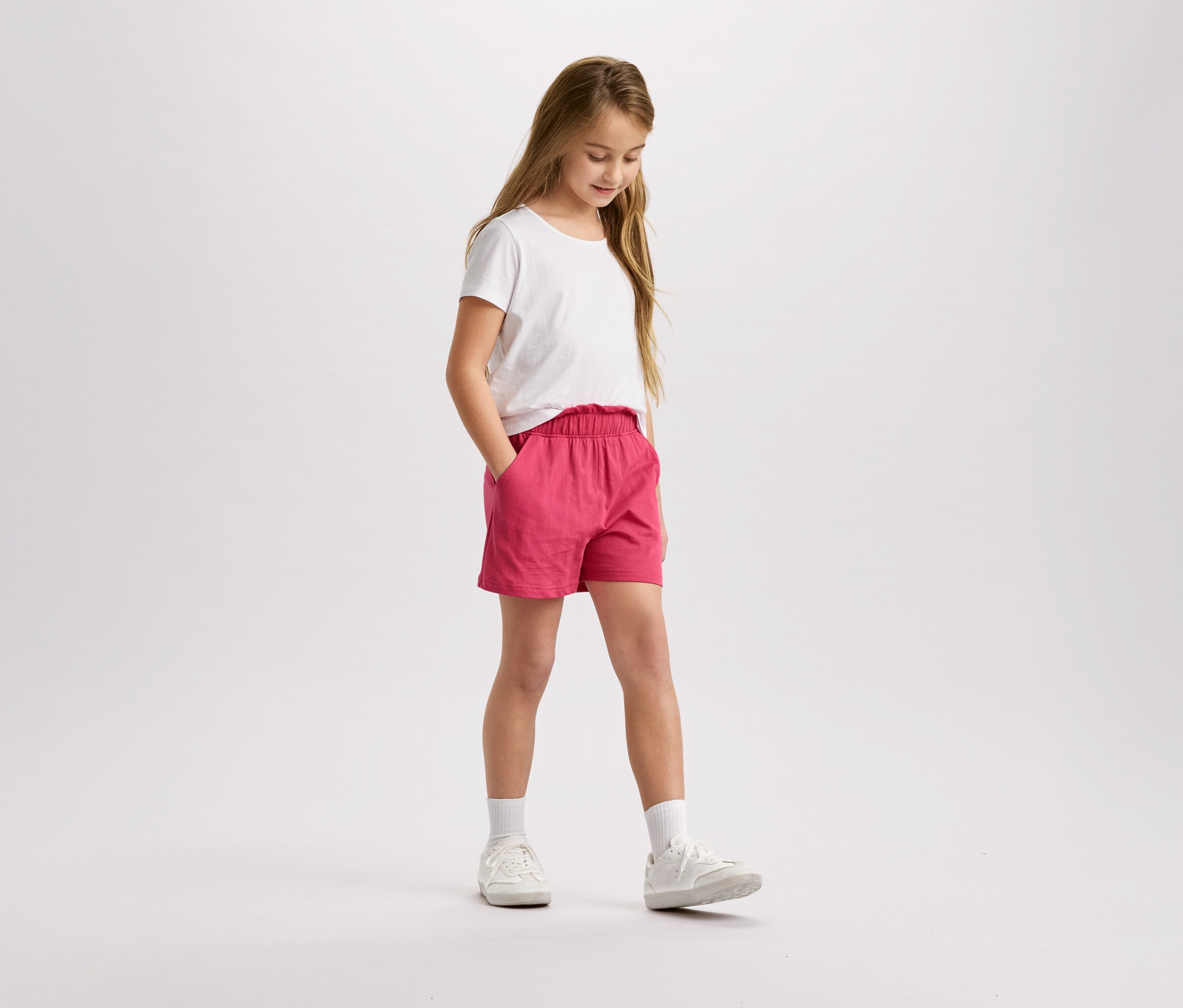 Dziewczyna z blond włosami w białej koszulce i różowych szortach stoi i patrzy w dół.