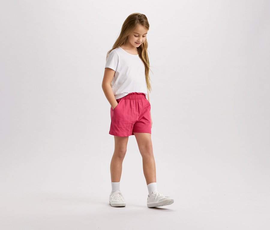 Dziewczyna z blond włosami w białej koszulce i różowych szortach stoi i patrzy w dół.