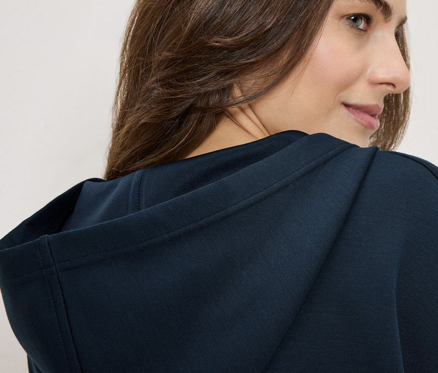 Kobieta ma na sobie granatową bluzę z kapturem, patrzy przez ramię.