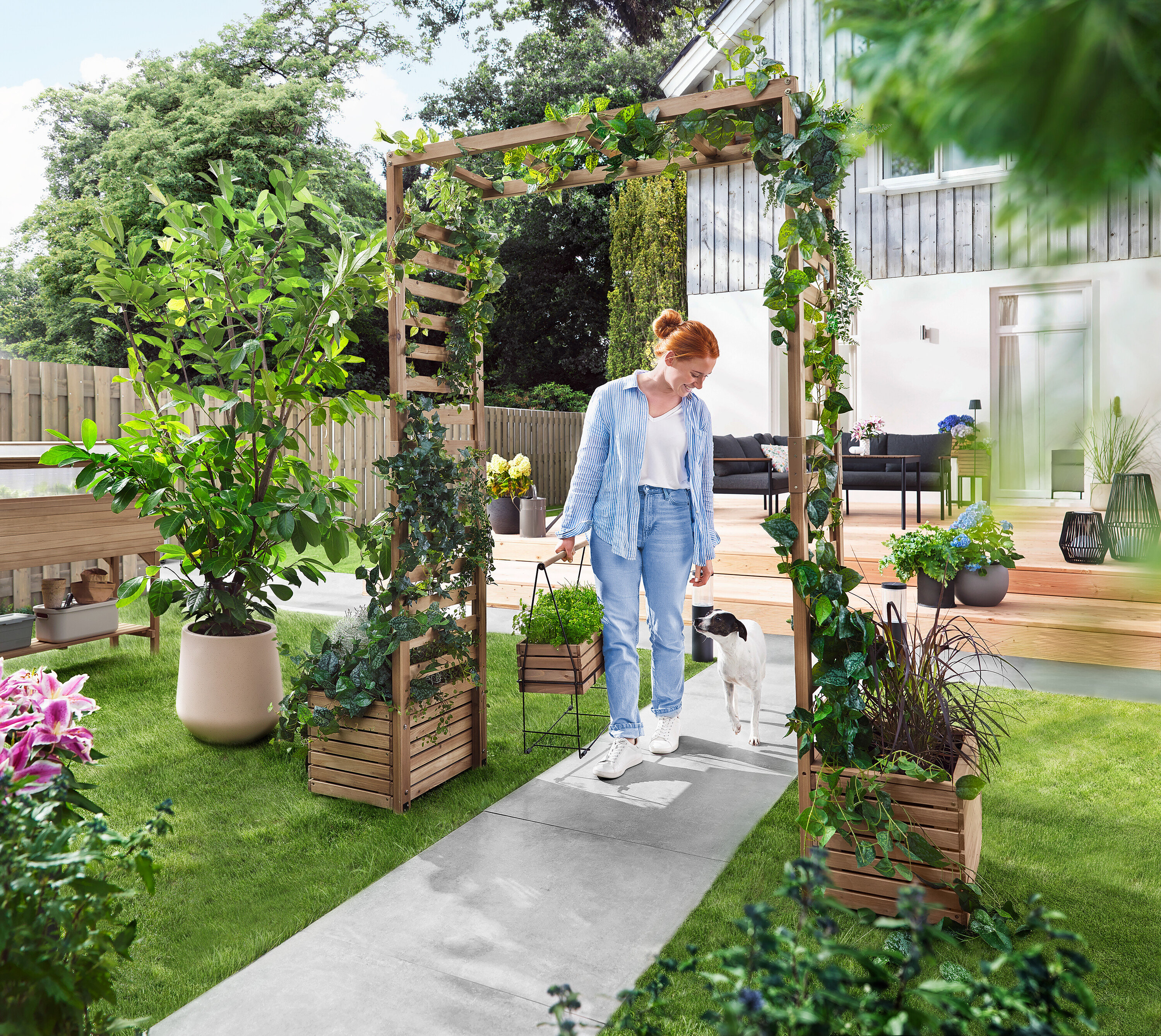 Kobieta idzie z psem przez łuk ogrodowy porośnięty roślinami. Niesie skrzynkę na rośliny.