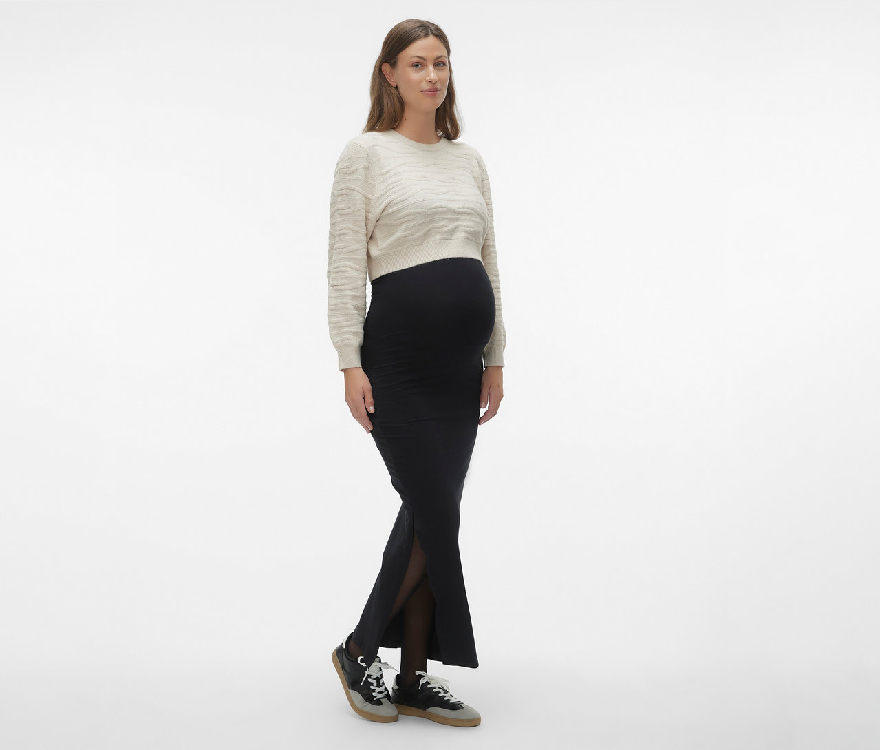 Kobieta ma na sobie czarną spódnicę maxi MAMALICIOUS „MLEMMA TUBE”, jasny sweter i trampki.