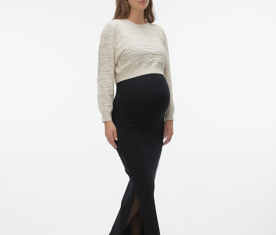 Kobieta w ciąży w beżowym swetrze i czarnej spódnicy z rozcięciem, do tego czarne buty.