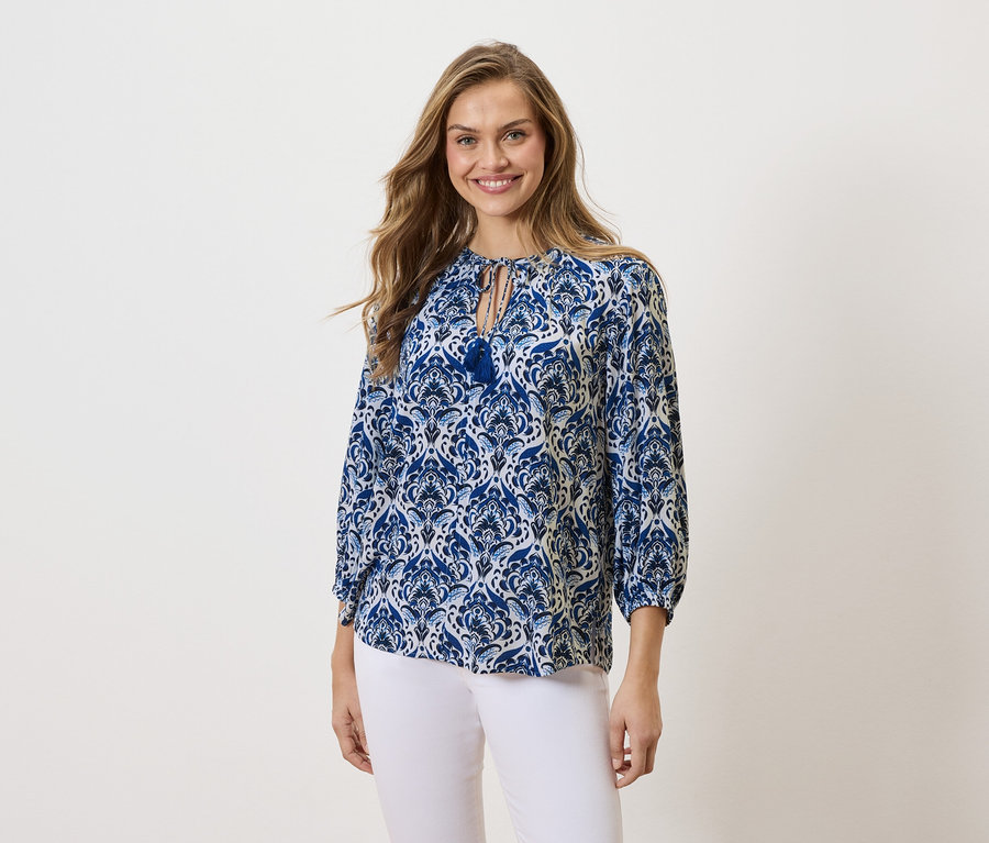 Kobieta z długimi blond włosami uśmiecha się, ubrana w bluzkę w niebiesko-biały wzór i białe spodnie.