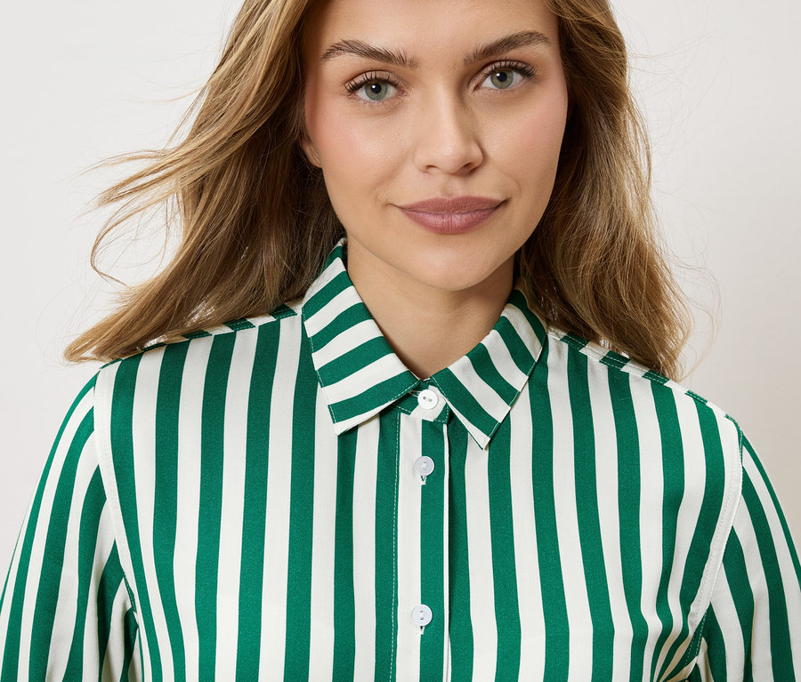 Portret kobiety o blond, falowanych włosach, ubranej w koszulę w paski w kolorze szmaragdowej zieleni i bieli.