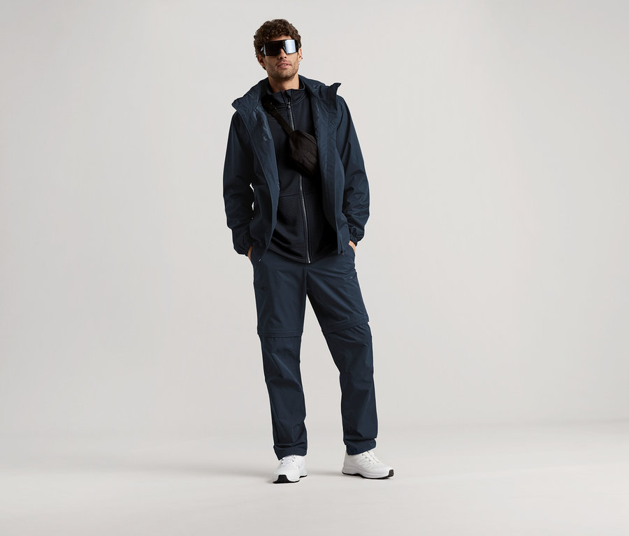 Mężczyzna stoi w okularach przeciwsłonecznych, granatowej kurtce, spodniach i torbie na biodra na jasnym tle.