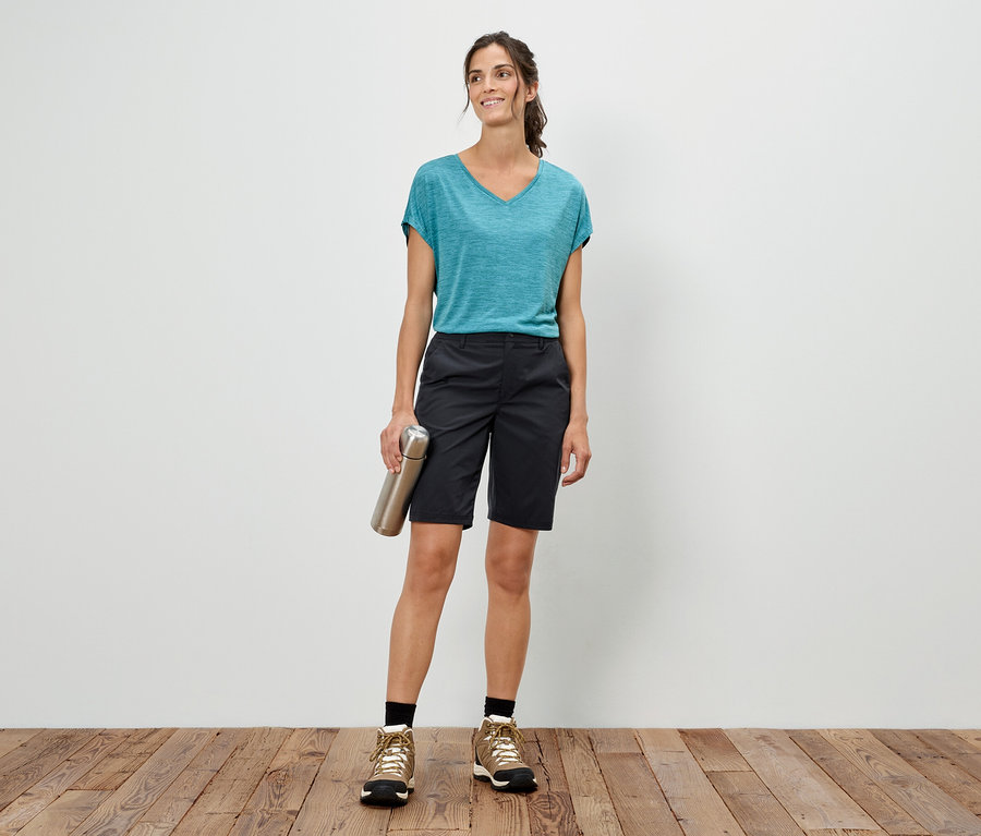 Kobieta stoi na tle białej ściany, trzyma srebrną butelkę, ma na sobie niebieski T-shirt, szorty i buty trekkingowe.