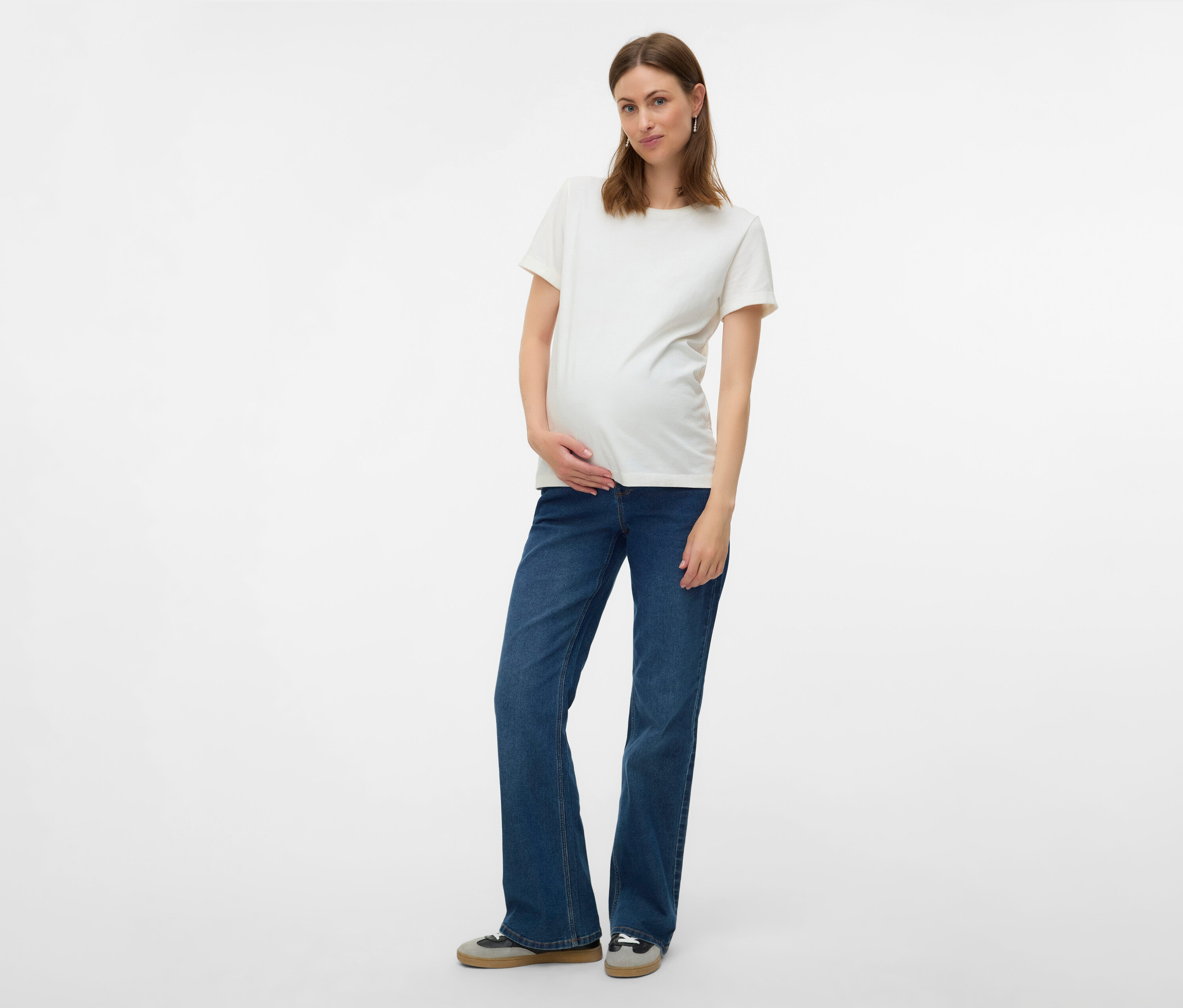 Kobieta w ciąży w białym T-shircie i rozszerzanych jeansach MAMALICIOUS „MLEDINA” pozuje na białym tle.