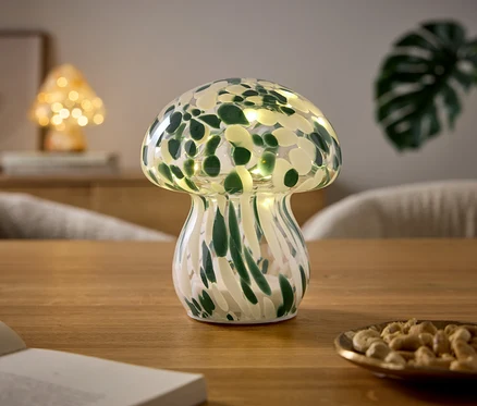 Lampa w kształcie grzyba z zielono-białym wzorem i światełkami stoi na drewnianym stole. Obok leży otwarta książka i mała miseczka z orzechami nerkowca. W tle widać kolejną lampę-grzyba i roślinę.