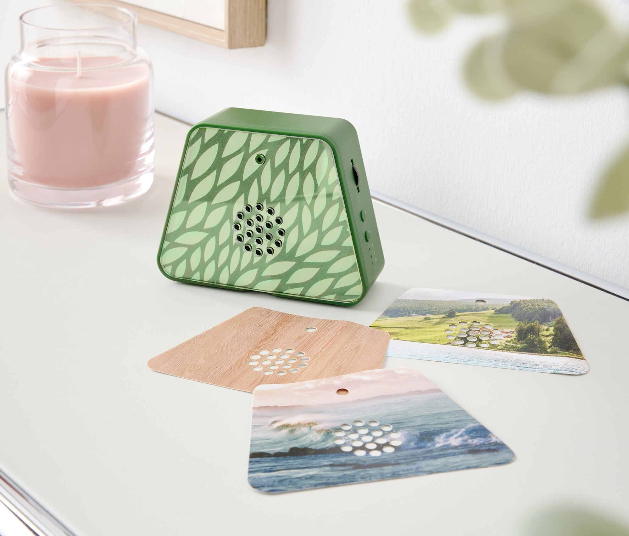 Na stole stoi różowa świeca, zielony dyfuzor zapachowy we wzór liści i trzy karty zapachowe.