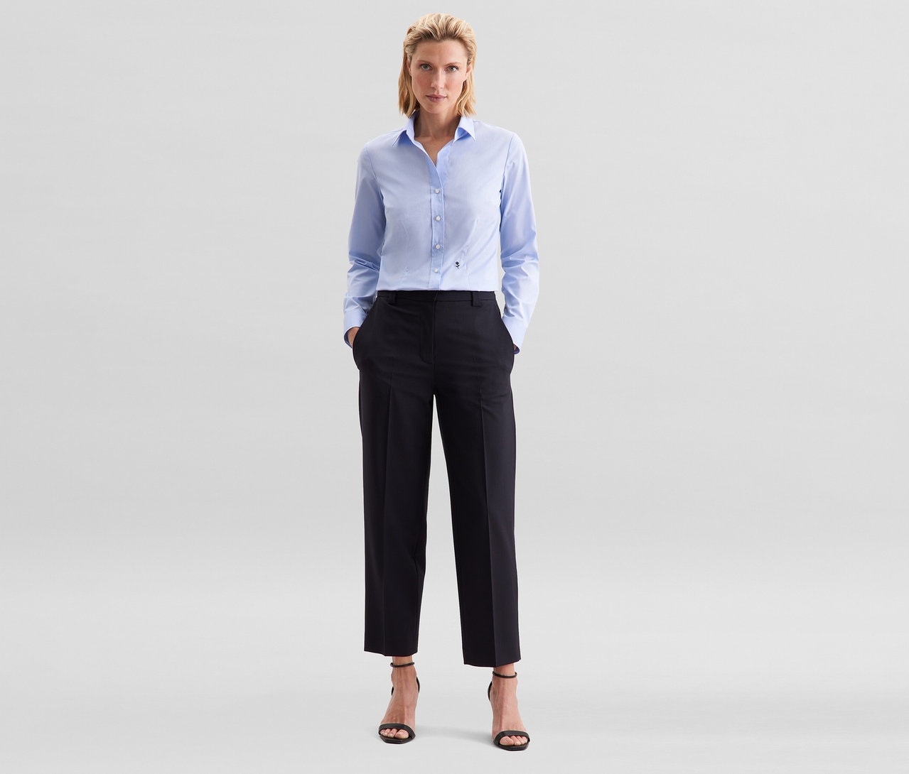 Kobieta stoi z rękami w kieszeniach, ma na sobie jasnoniebieską bluzkę Seidensticker Slim Fit non iron, czarne spodnie i czarne sandały na obcasie.