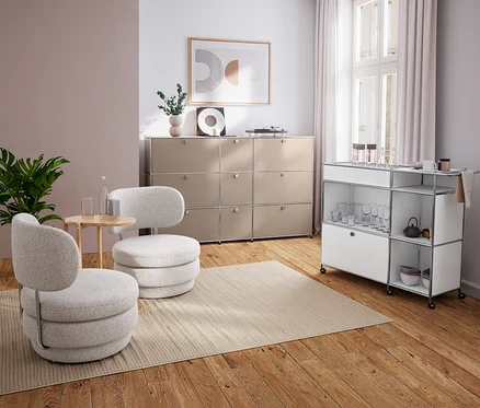 Nowoczesny salon z dwoma jasnoszarymi fotelami, okrągłym stolikiem z litego drewna, beżową komodą i białym wózkiem barowym.