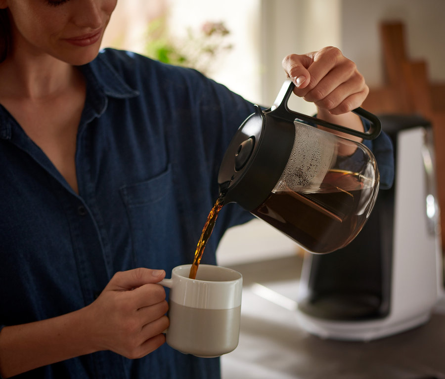 Kobieta nalewa kawę z dzbanka do filiżanki. W tle widoczny ekspres przelewowy »Let's Brew«.