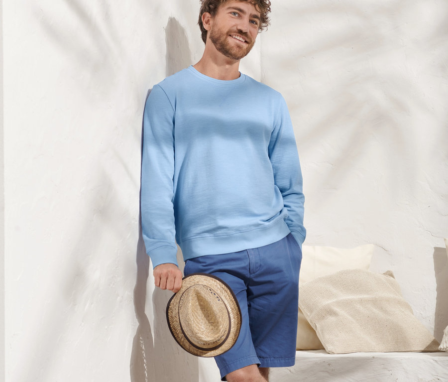 Mężczyzna oparty o ścianę trzyma kapelusz i ma na sobie jasnoniebieską bluzę.