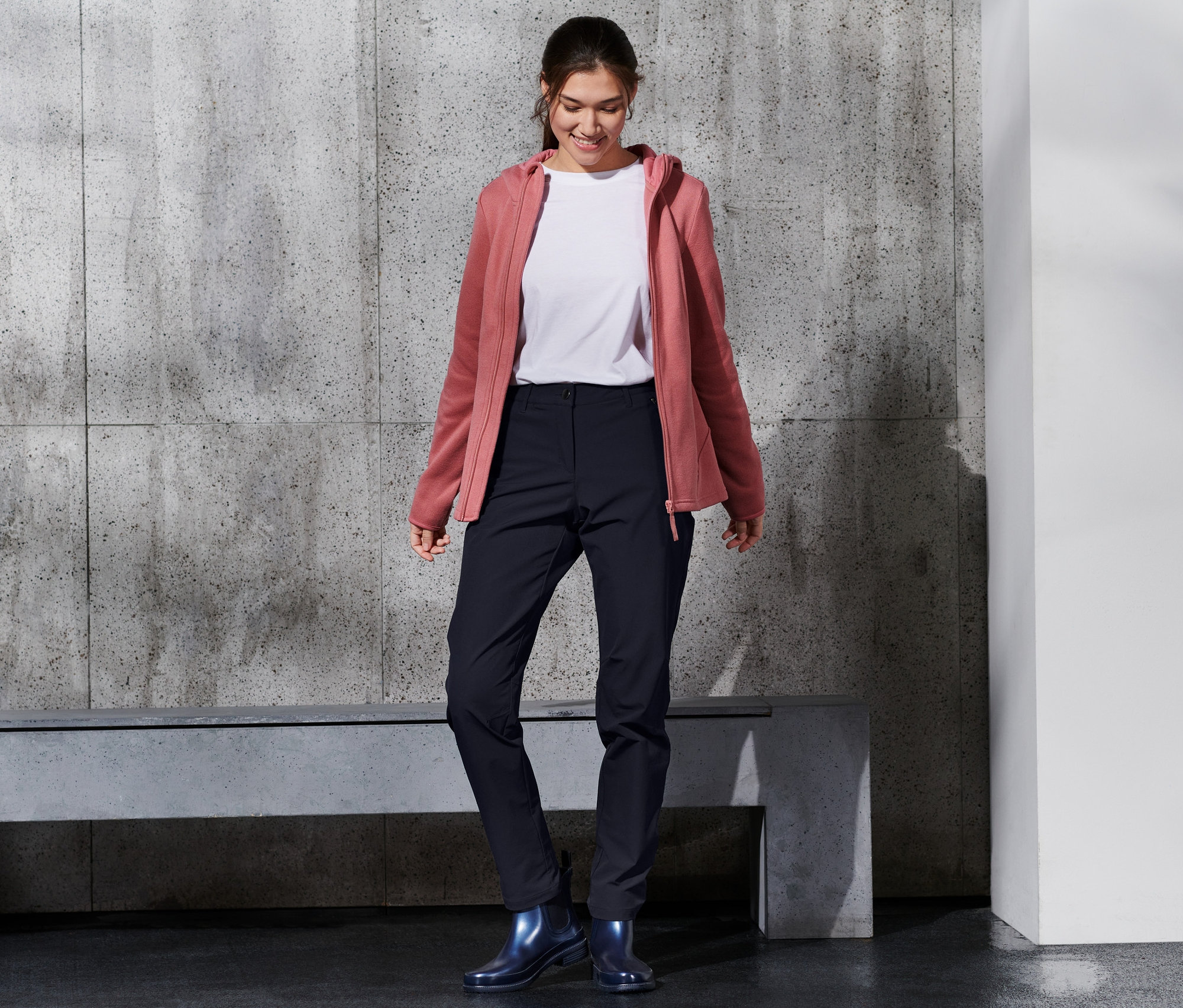 Uśmiechnięta kobieta ma na sobie spodnie z softshellu, biały t-shirt i różową bluzę z kapturem.