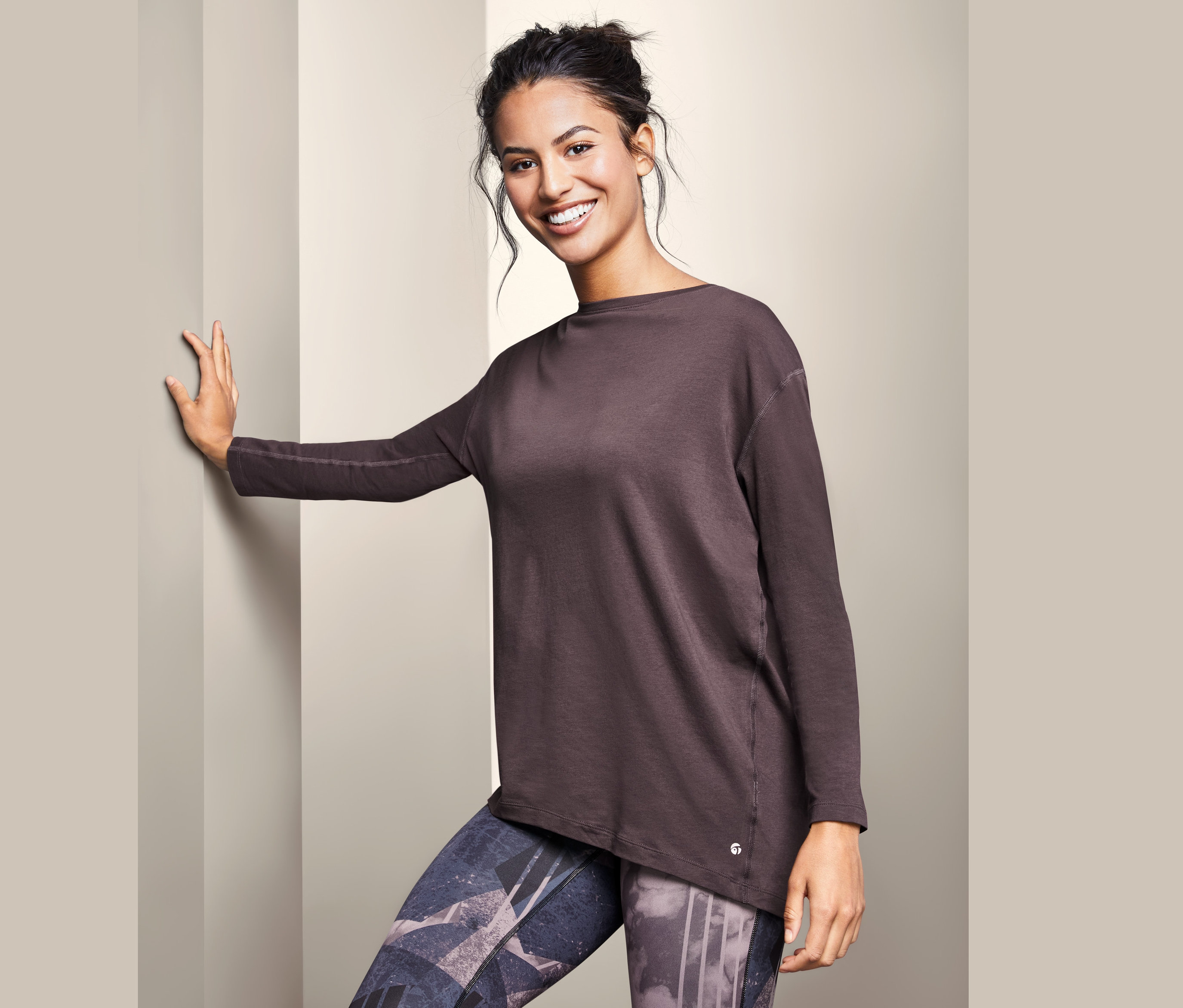 Kobieta oparta o ścianę ma na sobie długą koszulkę funkcyjną i wzorzyste legginsy.