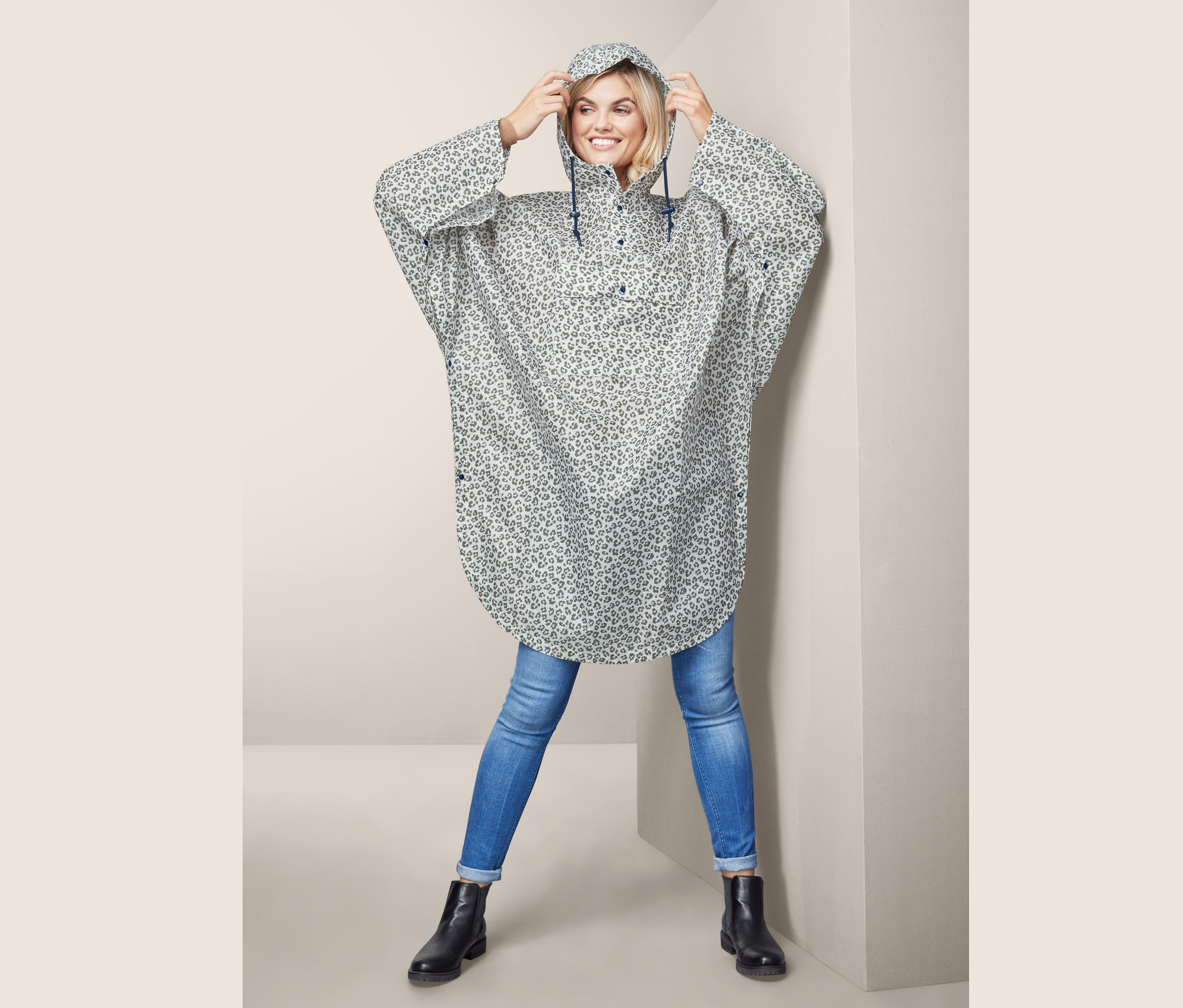 Kobieta poprawia kaptur poncza przeciwdeszczowego w lamparcie wzory. Ma na sobie dżinsy i czarne buty.