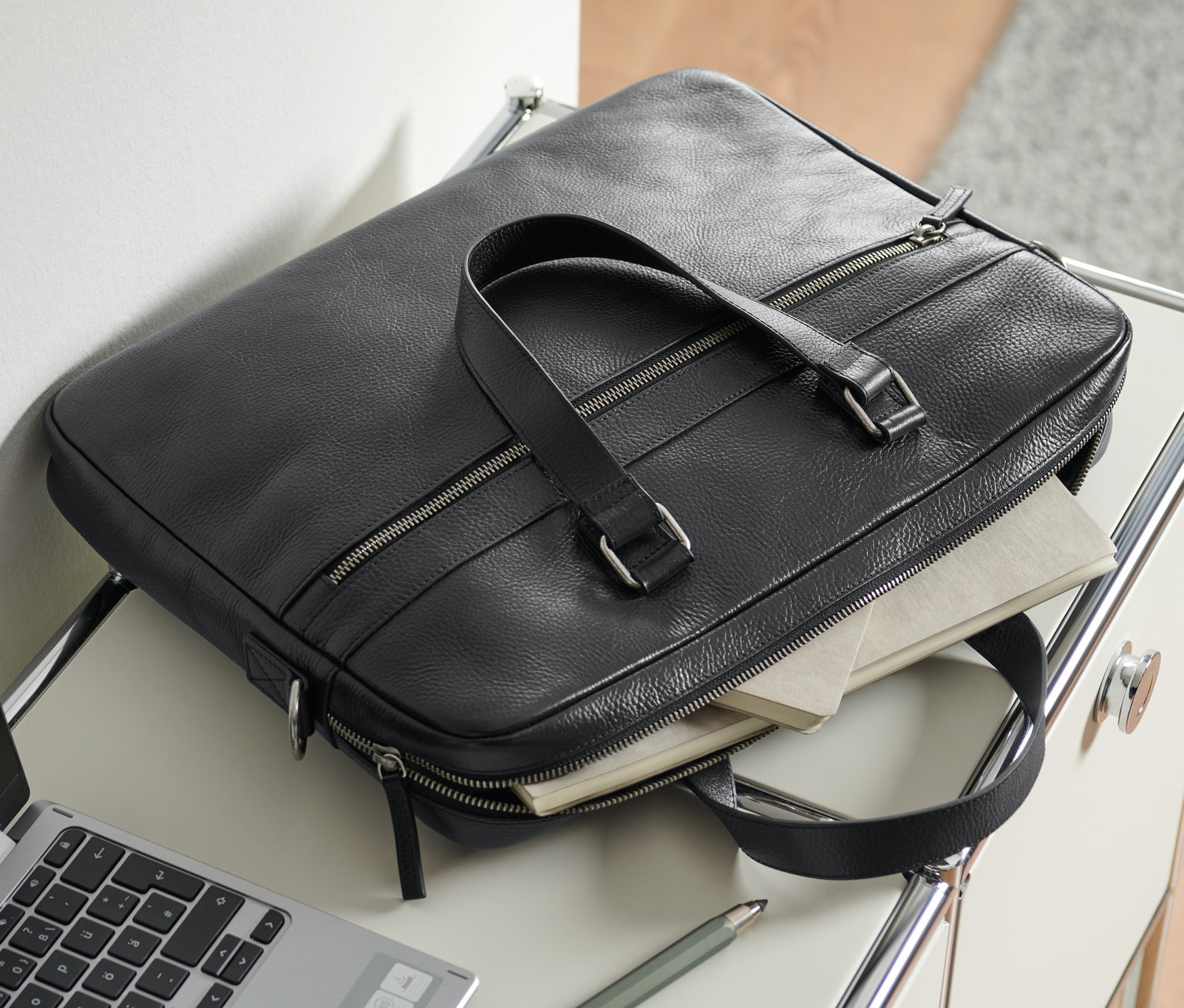 Czarna skórzana aktówka leży na stole obok laptopa i długopisu.