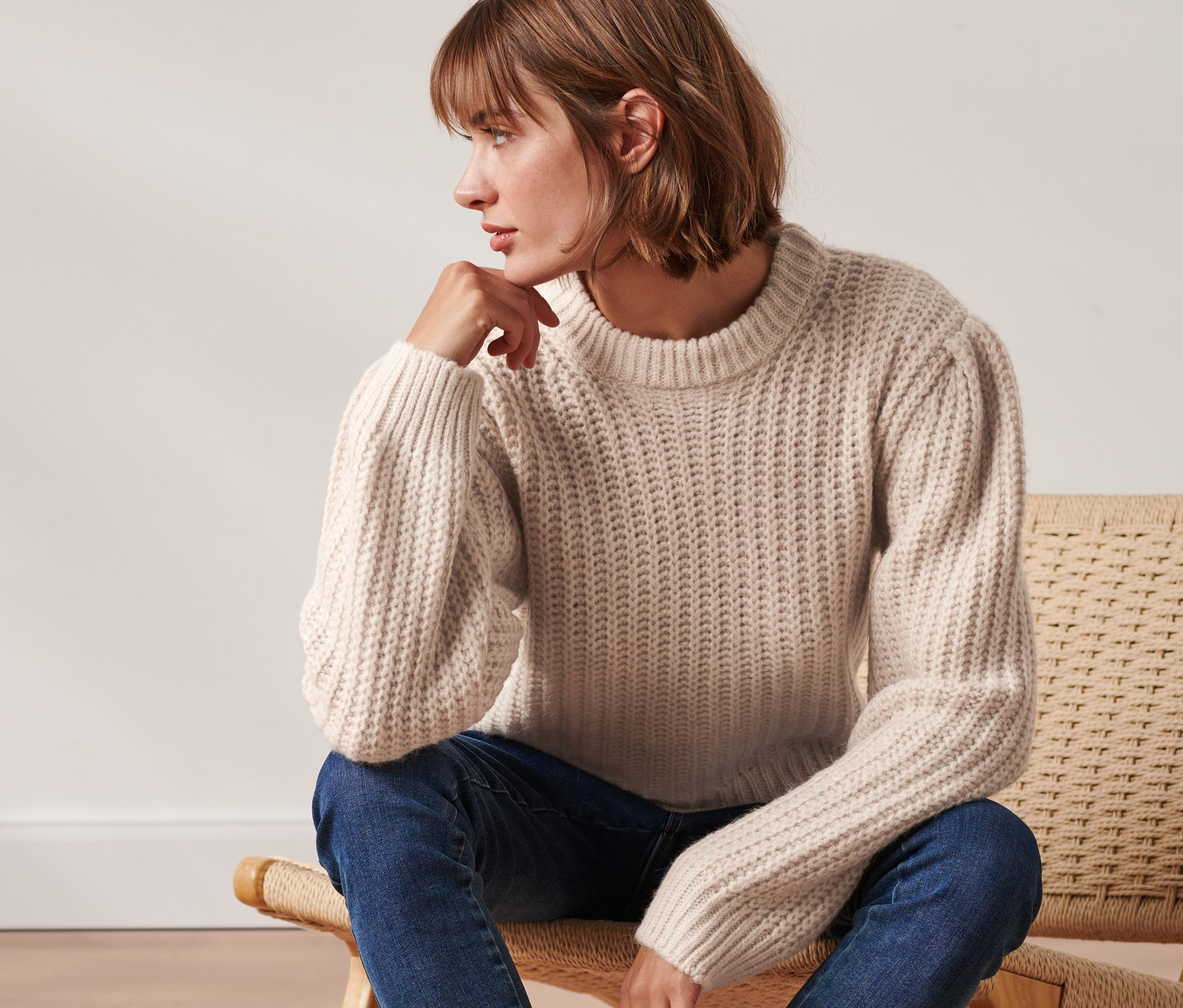 Kobieta siedzi na krześle i ma na sobie kremowy sweter z dzianiny o luźnym splocie, z wełną, niebieskie dżinsy i czarne buty.