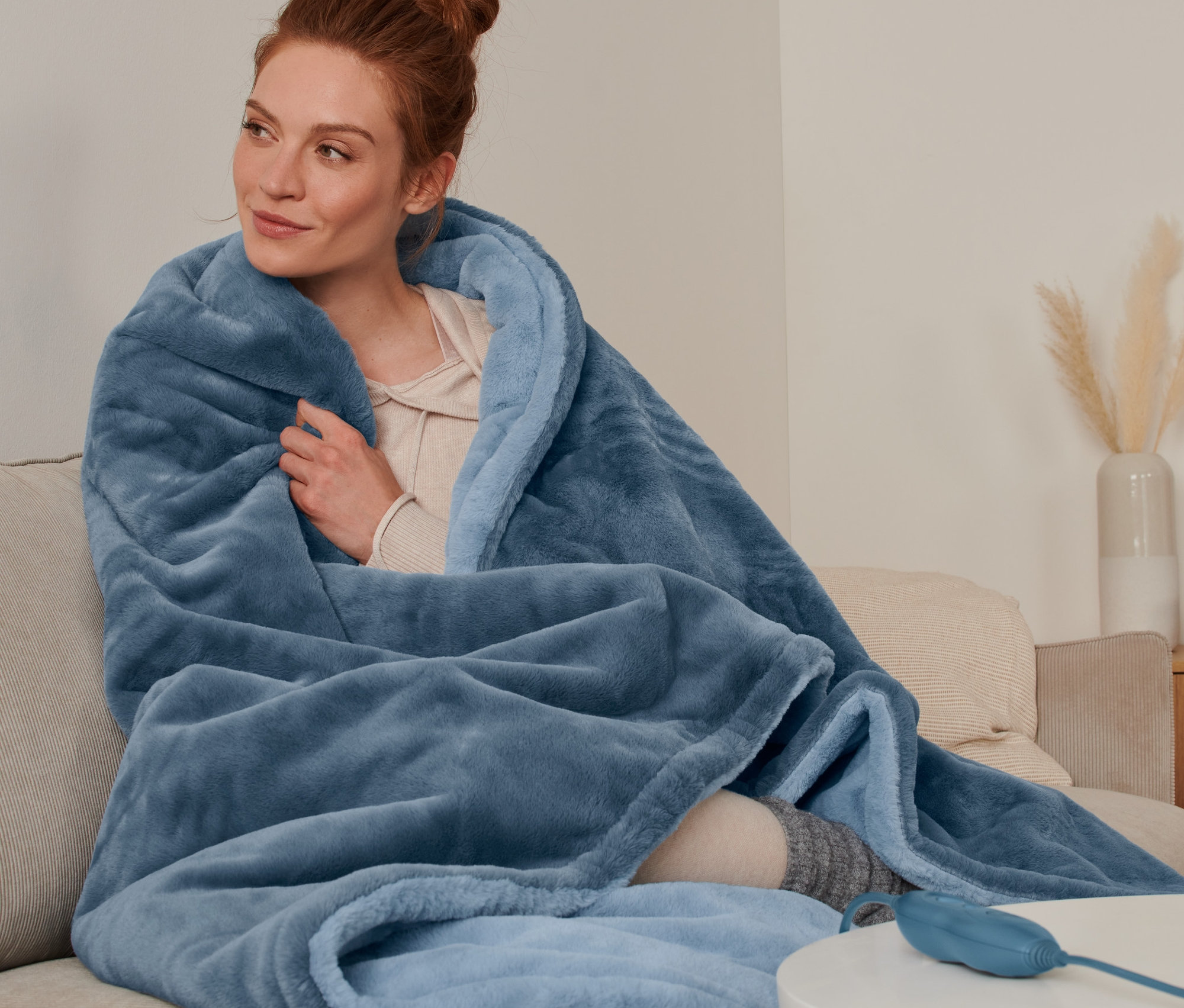 Kobieta siedzi na sofie owinięta kocem elektrycznym.