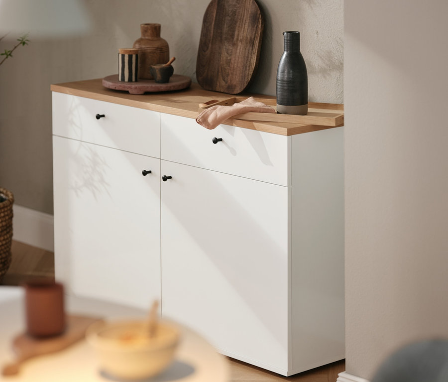 Na białej komodzie stoi brązowa deska do krojenia i różne przedmioty dekoracyjne. Nad nią wisi lampa sufitowa »Strap 16« Design for the people by Nordlux.