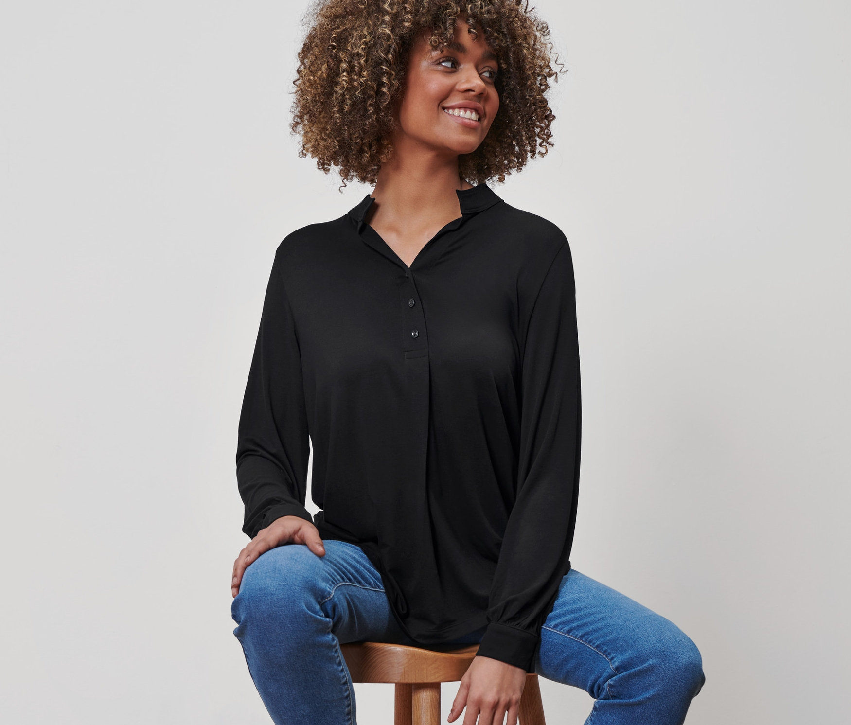 Kobieta siedzi na stołku ubrana w czarną bluzkę z plisą z guzikami, niebieskie dżinsy i czarne mokasyny.