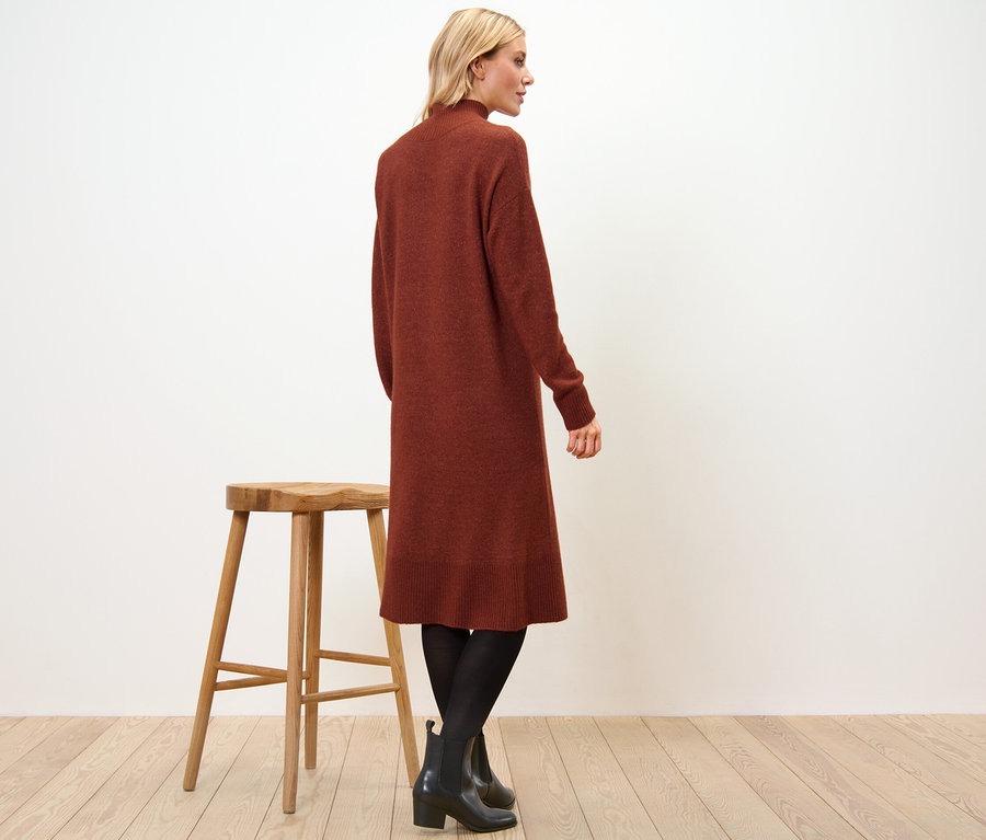 Kobieta stoi bokiem, ubrana w dzianinową sukienkę z zawartością wełny, czarne rajstopy i botki. Obok niej stoi drewniany stołek.