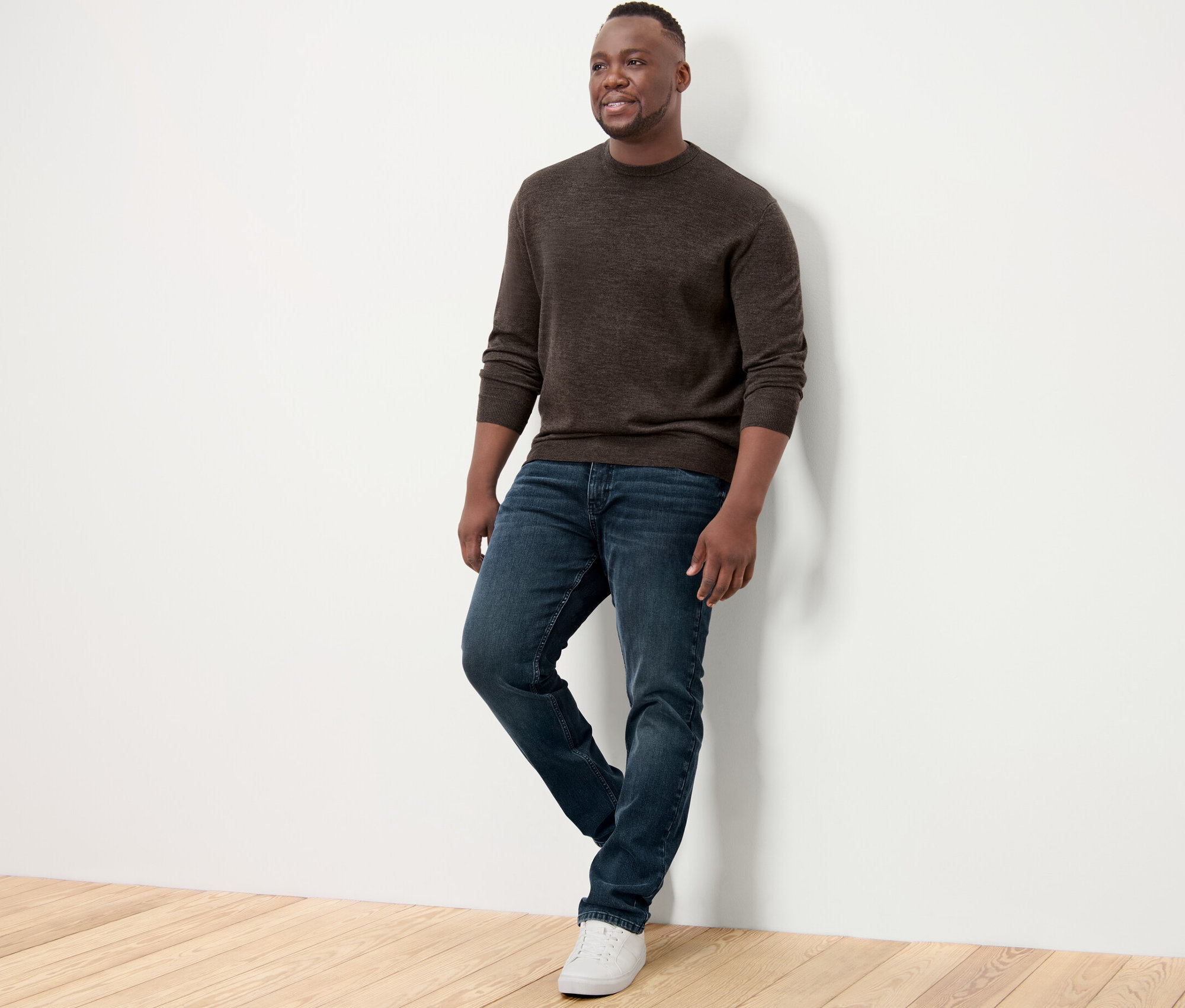 Mężczyzna oparty o ścianę ma na sobie brązowy sweter z wełny merino, dżinsy i tenisówki.