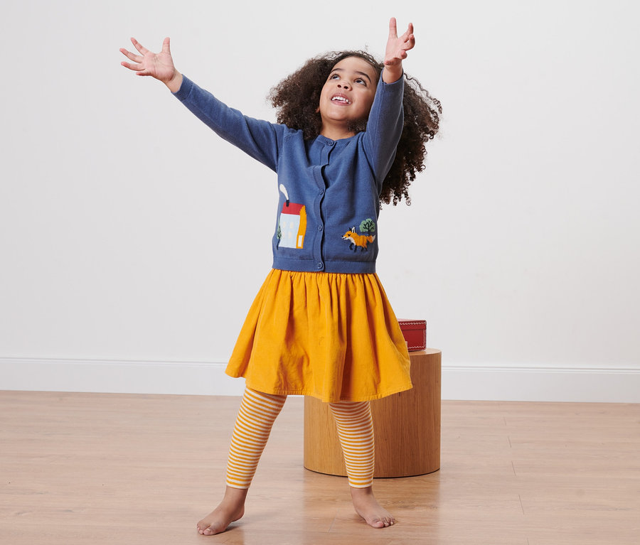 Dziewczynka z kręconymi włosami stoi z podniesionymi rękami. Ma na sobie niebieski sweter z aplikacjami, żółtą spódnicę i dziecięce legginsy termoaktywne, 3 pary, paski.