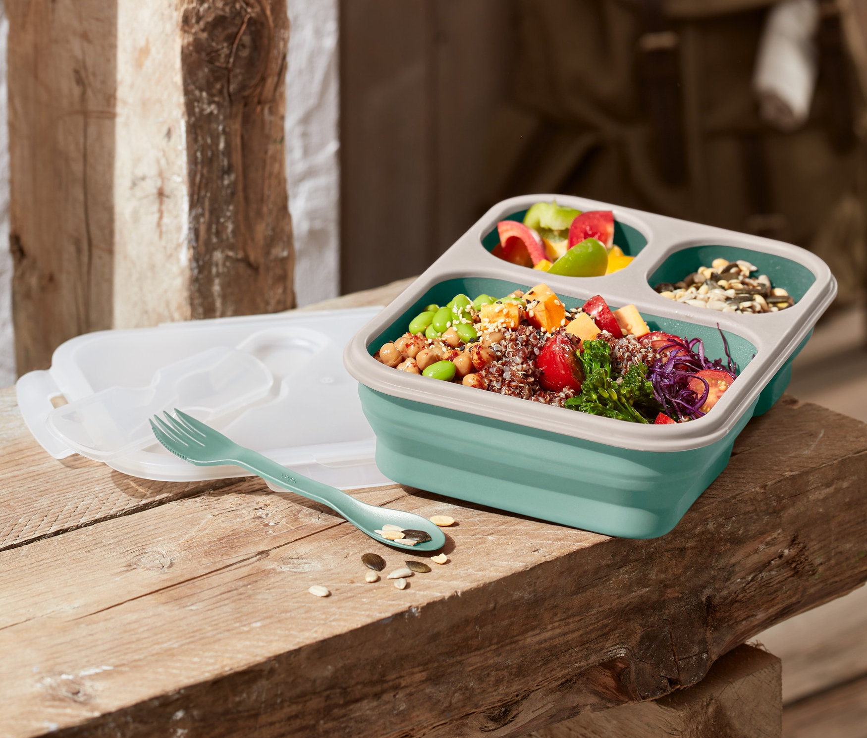 Składany pojemnik na jedzenie stoi na drewnianej ławce, obok leżą widelec i pokrywka.
