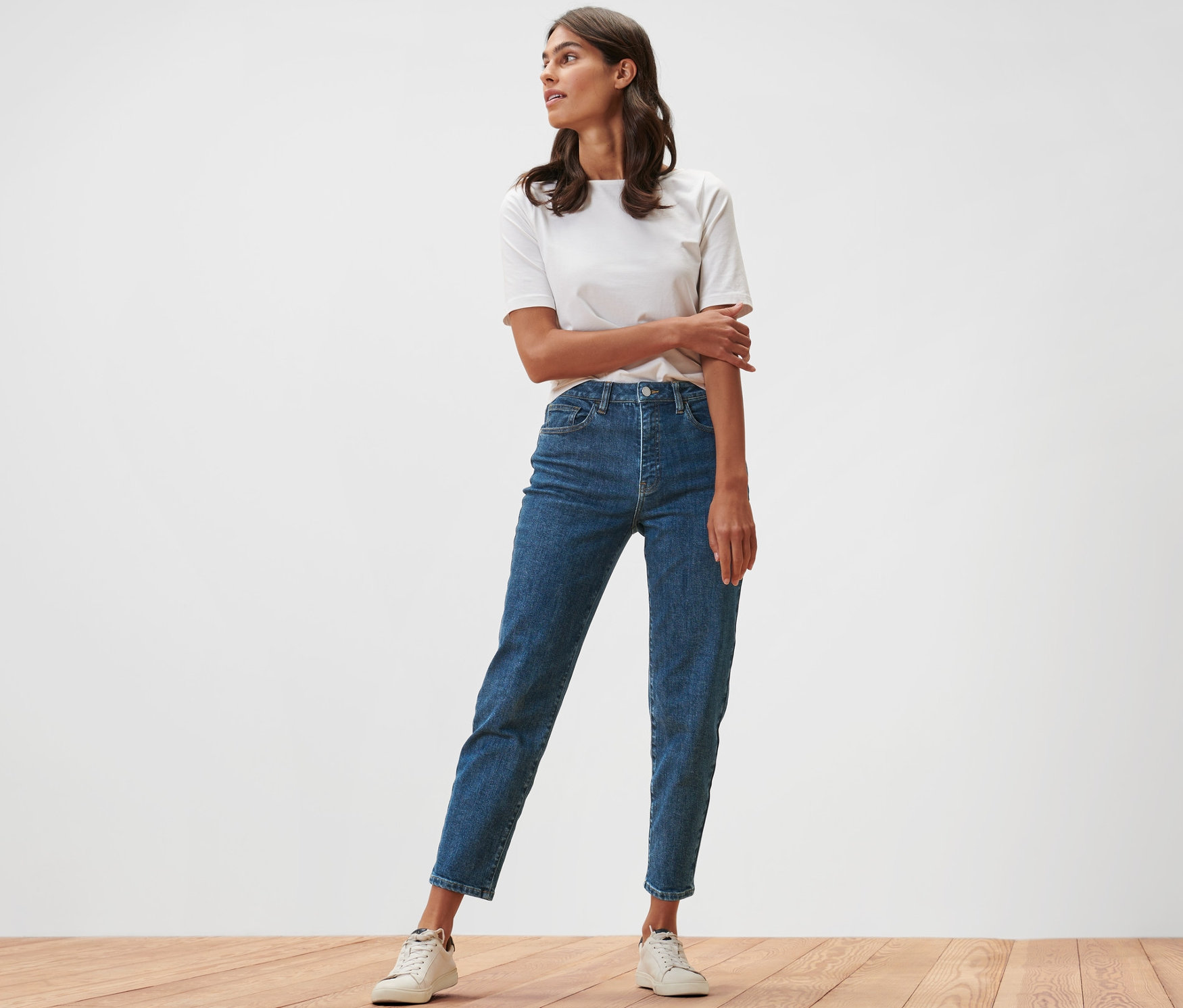 Kobieta stoi na drewnianej podłodze w dżinsach typu Mom Jeans – fason „Nyla” i białym T-shircie.