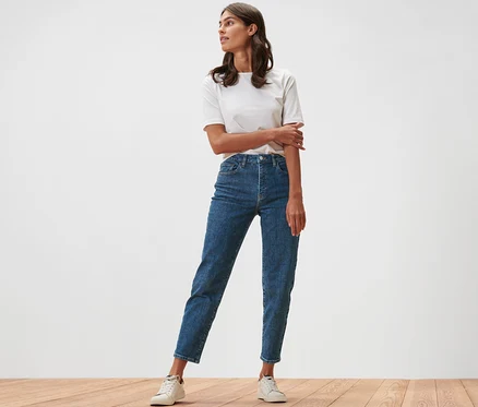 Kobieta stoi na drewnianej podłodze w dżinsach typu Mom Jeans – fason „Nyla” i białym T-shircie.