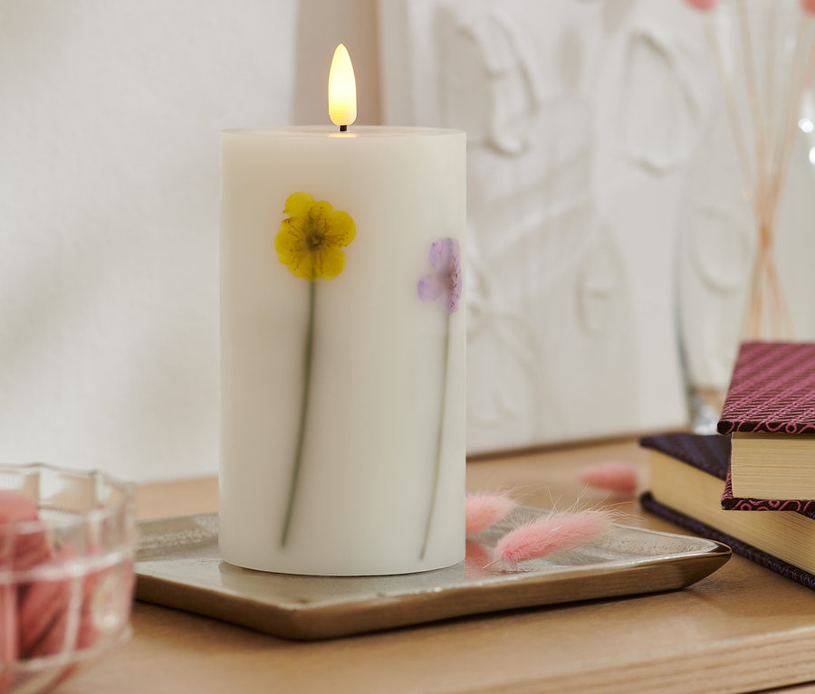 Na tacy stoi świeca LED z prawdziwego wosku z suszonymi kwiatami. W tle książki i wazon.