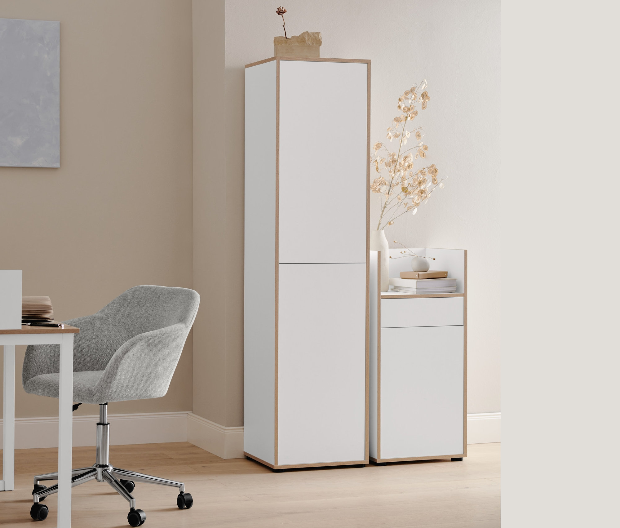 Beżowe krzesło biurowe stoi przy białym biurku z bocznym obramowaniem. Obok stoi wysoka i mała szafka biurowa z fornirowanymi krawędziami. Na stole stoi metalowa lampka stołowa.