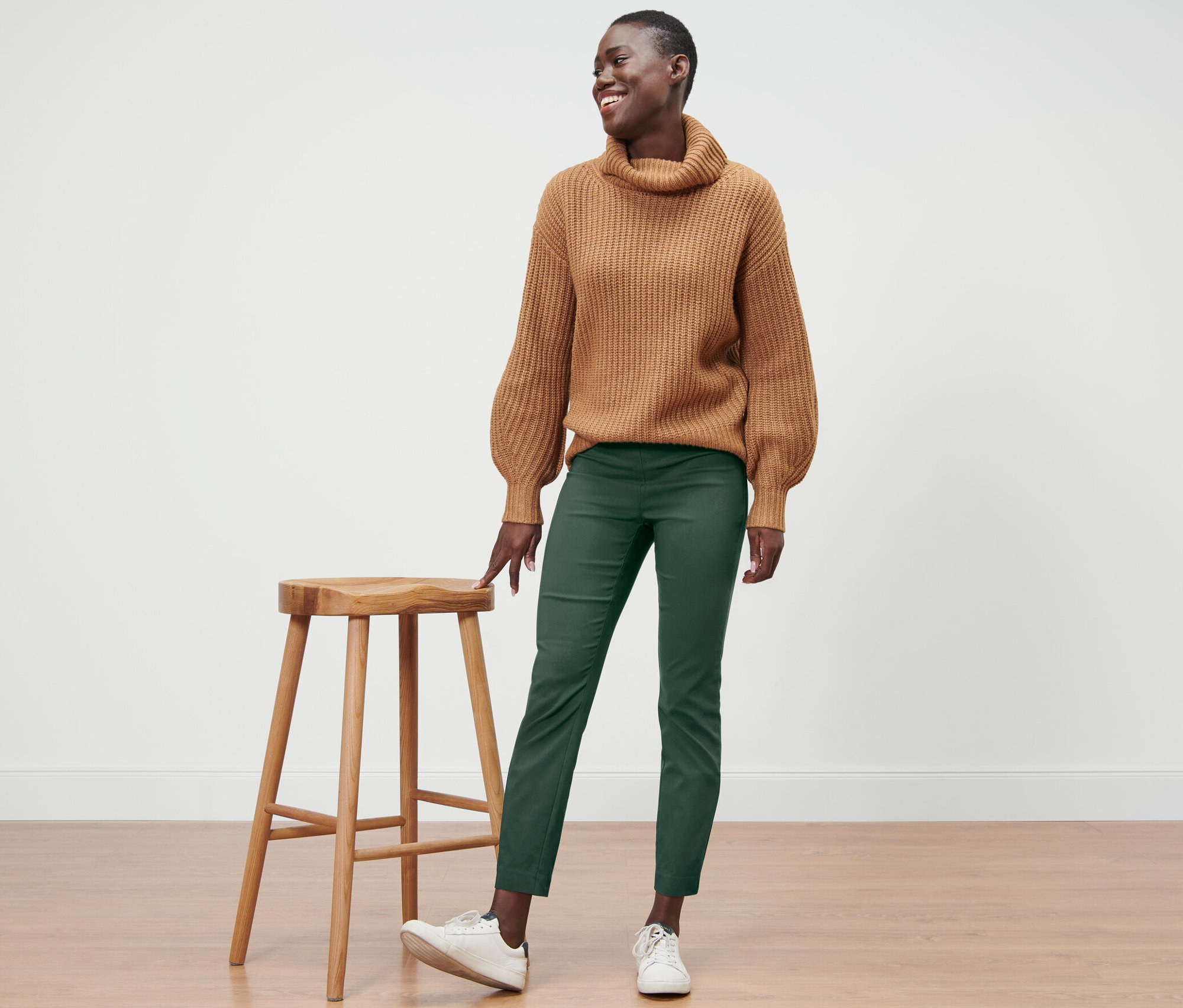 Uśmiechnięta kobieta stoi obok drewnianego stołka, ubrana w brązowy sweter i zielone spodnie stretchowe.