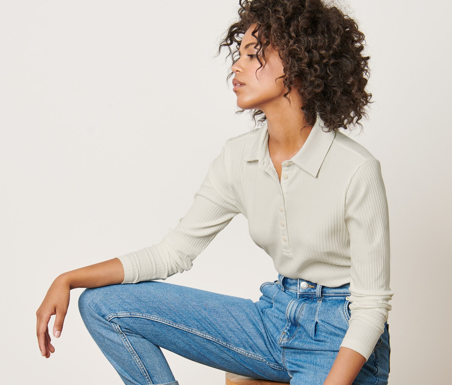 Kobieta siedzi na drewnianym stołku, ubrana w trykotową koszulkę z kołnierzykiem, dżinsy i tenisówki.